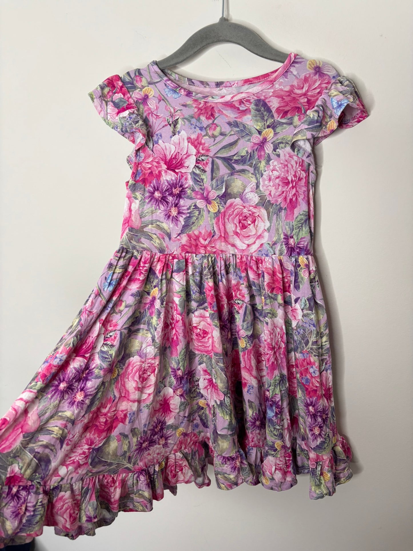 Pink and purple floral print dress with cap sleeves and ruffle hem, hanging on a hanger against a white brick wall.