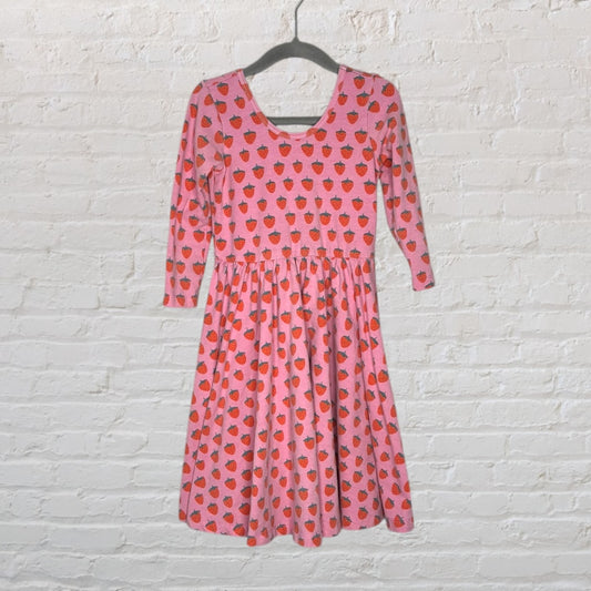 Pink skater dress with red strawberry print and three-quarter sleeves, hanging on a hanger against a white brick background.