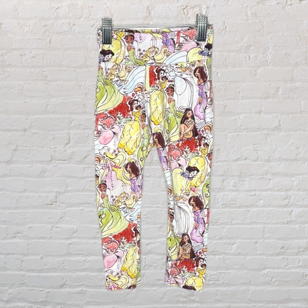 Multicolored leggings featuring various Disney princess characters, displayed against a white brick wall.