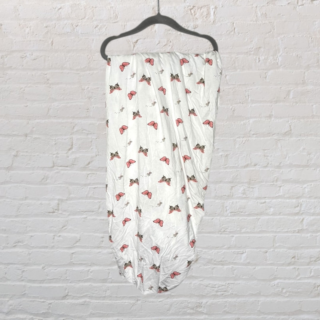 White cotton swaddle blanket with pink and brown butterfly prints hanging on a black hanger against a brick wall.