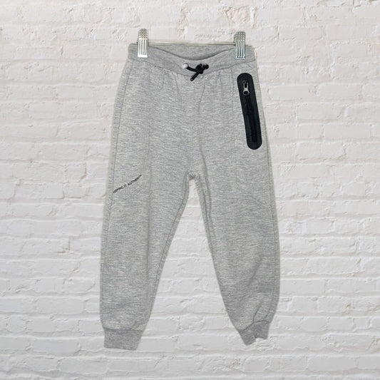 Grey jogger sweatpants with a black zipper pocket and drawstring, hanging on a white brick wall.