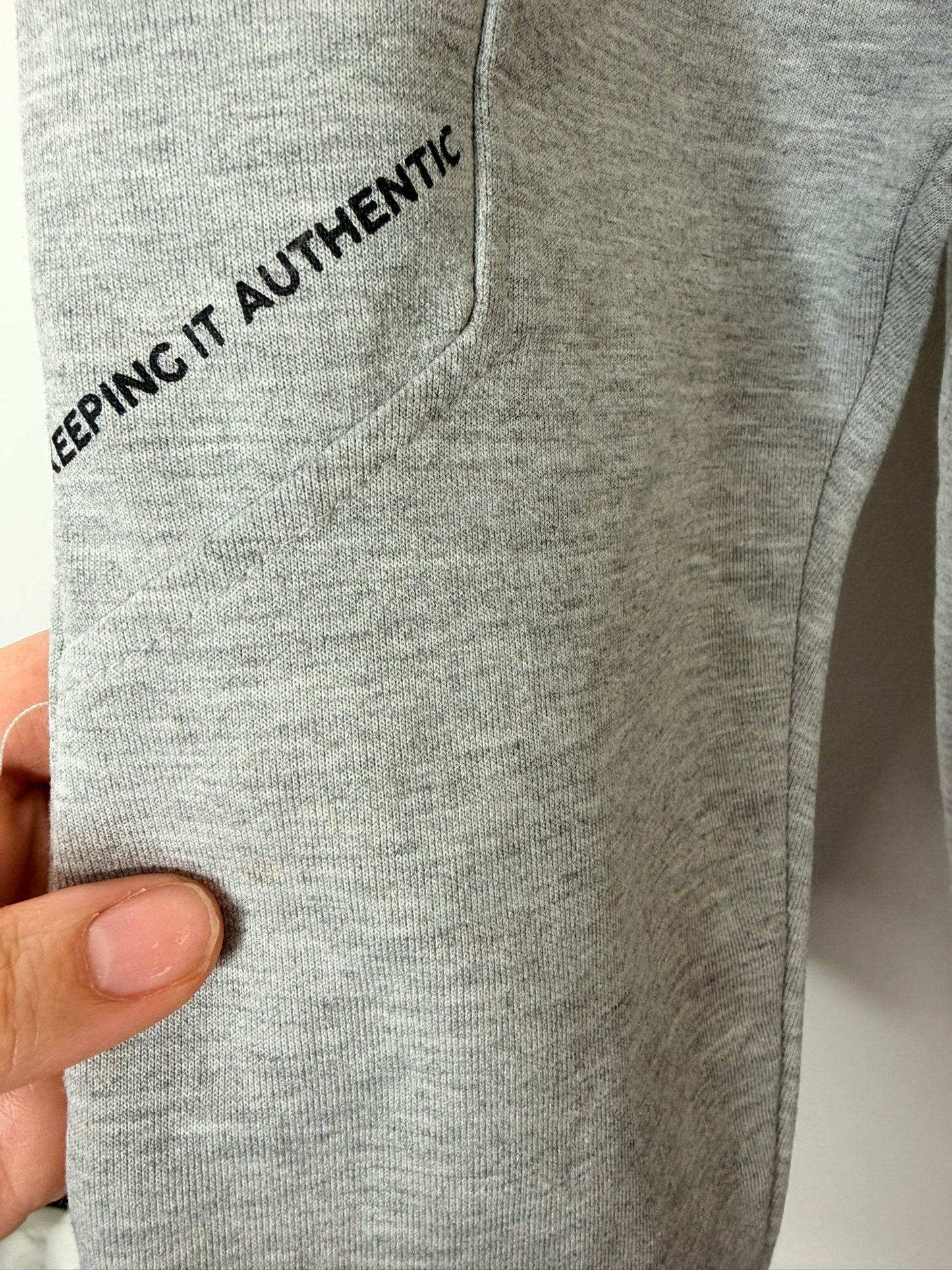 Grey jogger sweatpants with a black zipper pocket and drawstring, hanging on a white brick wall.