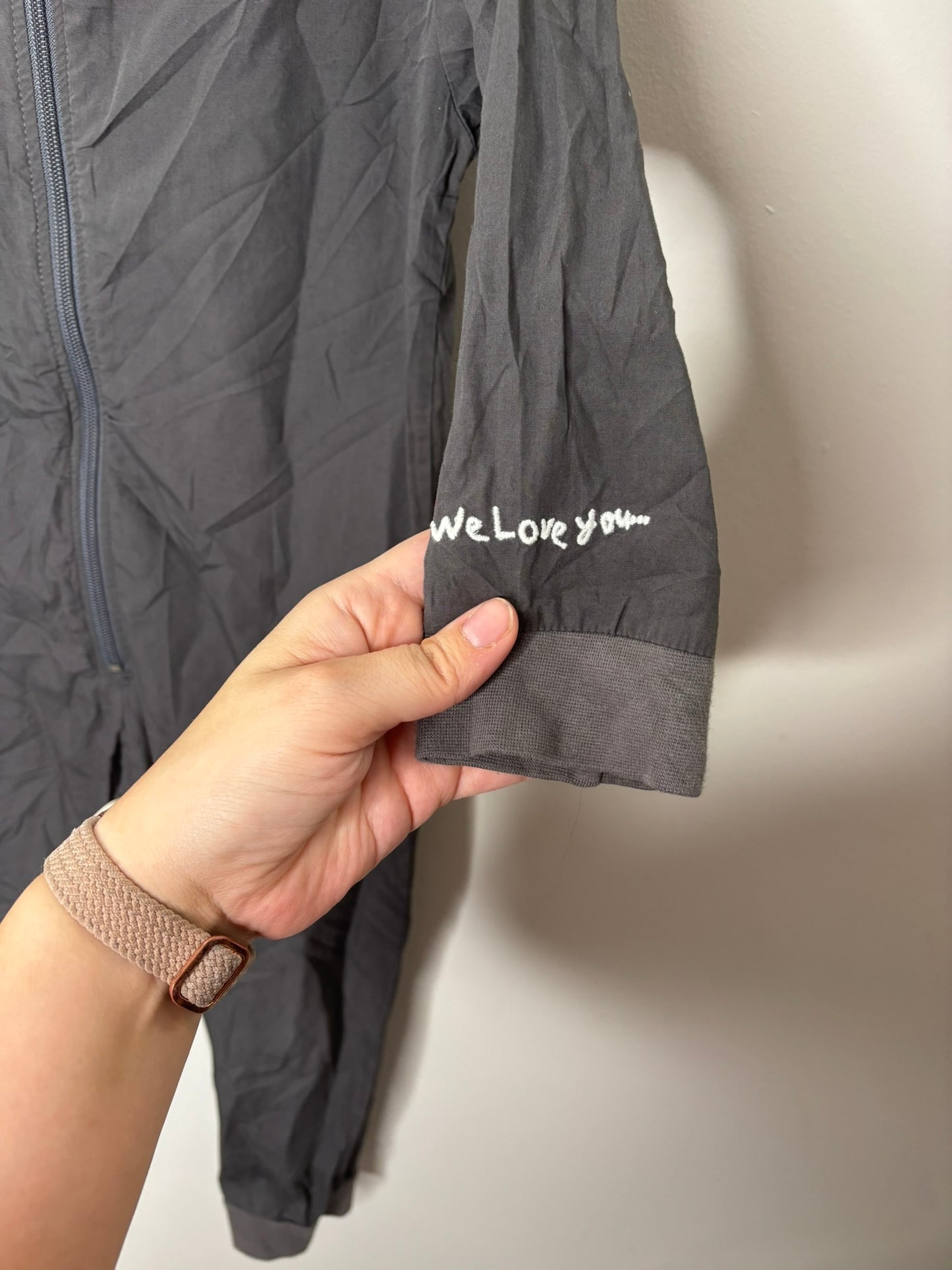 Dark grey coveralls with 'Hotel Cleaning Crew' printed on the back, displayed on a hanger against a white brick wall.