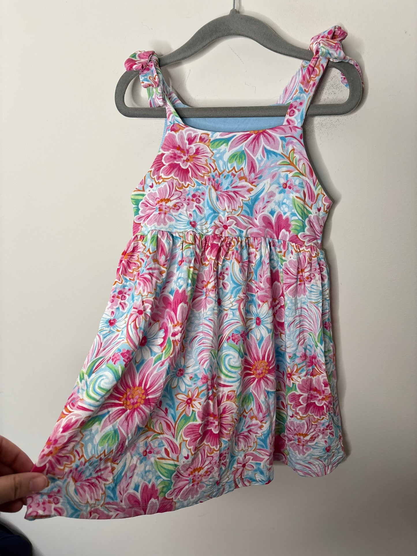 A floral sundress with ruffle straps featuring pink, blue, and green flowers, hanging on a gray hanger.