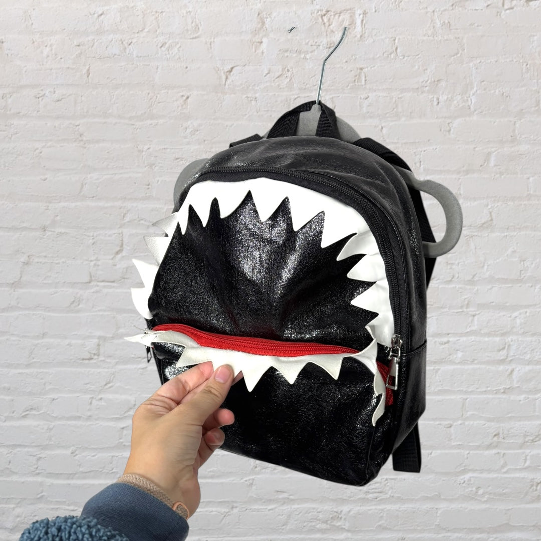 Black glittery shark-themed backpack with white jagged teeth design, hanging against a white brick wall.