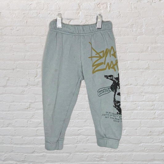 Gray jogger sweatpants with graphic print including text and imagery, displayed hanging on clips against a brick background.