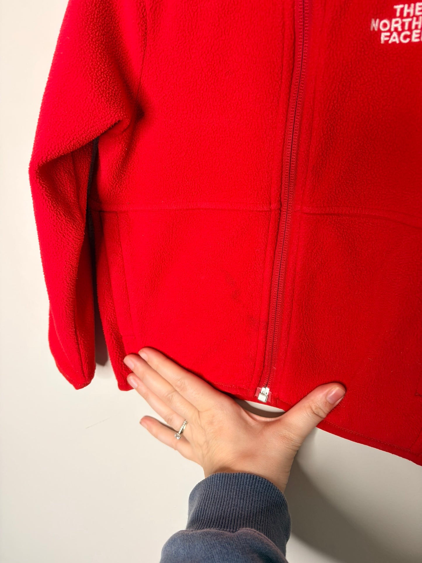Bright red fleece zip-up hoodie by The North Face with a hood, displayed against a white brick wall.