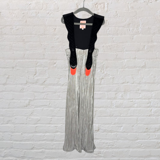Silver pleated overalls with black quilted suspenders and bright orange accents, hanging on a hanger against a white brick wall.