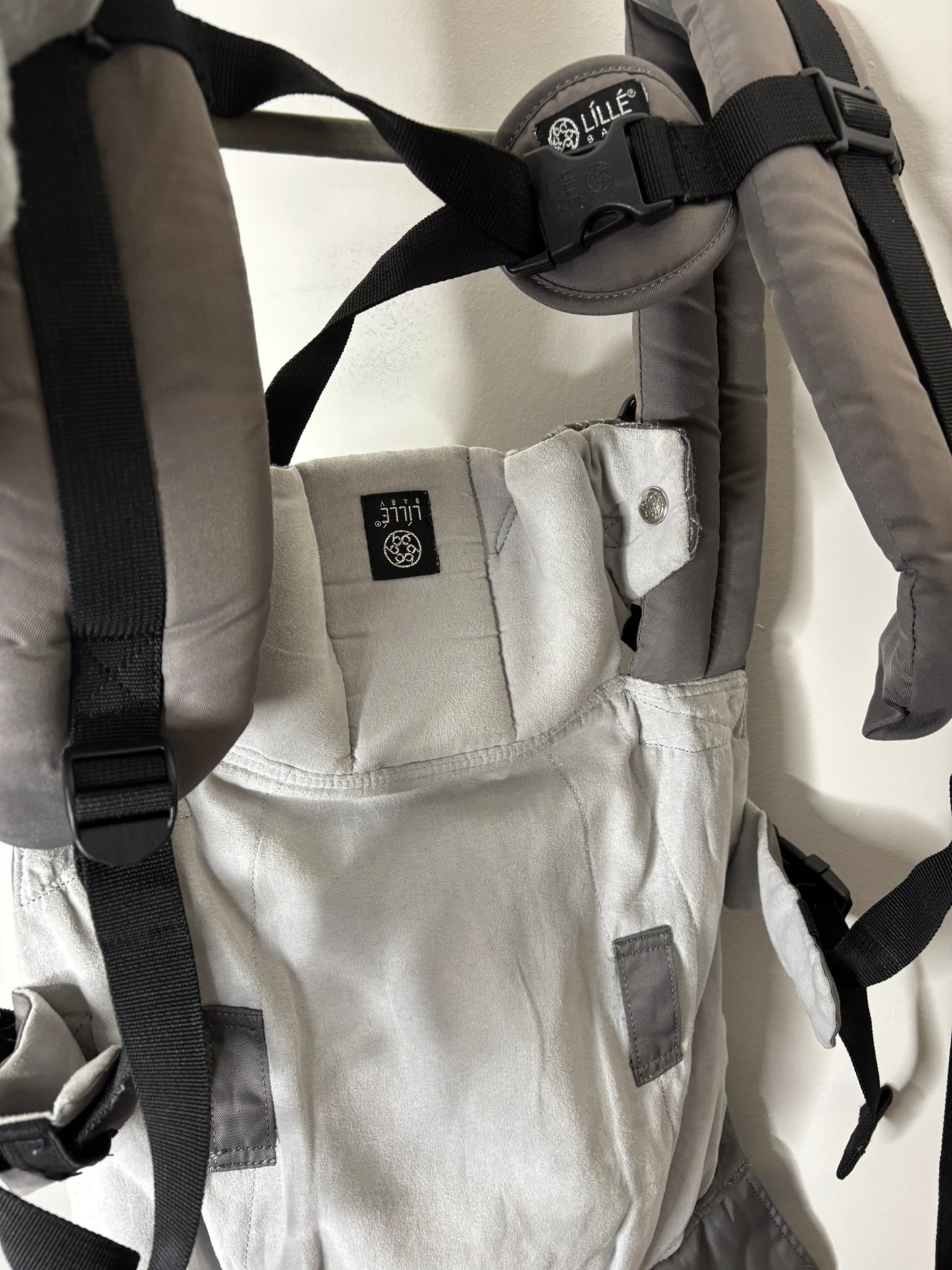 Gray ergonomic baby carrier with multiple straps and support, hanging on a hanger.
