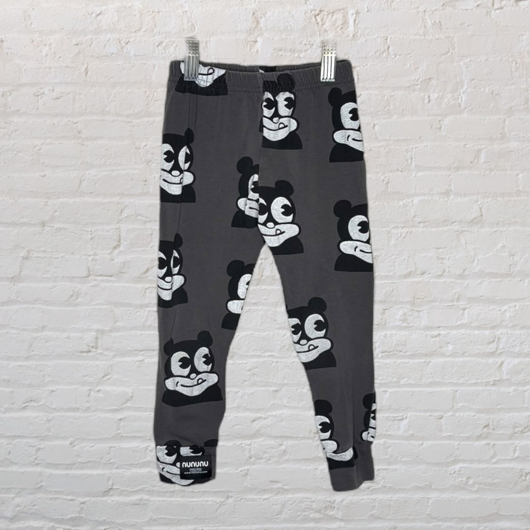 Dark grey leggings with white graphic face prints by Nununu, displayed against a white brick wall.
