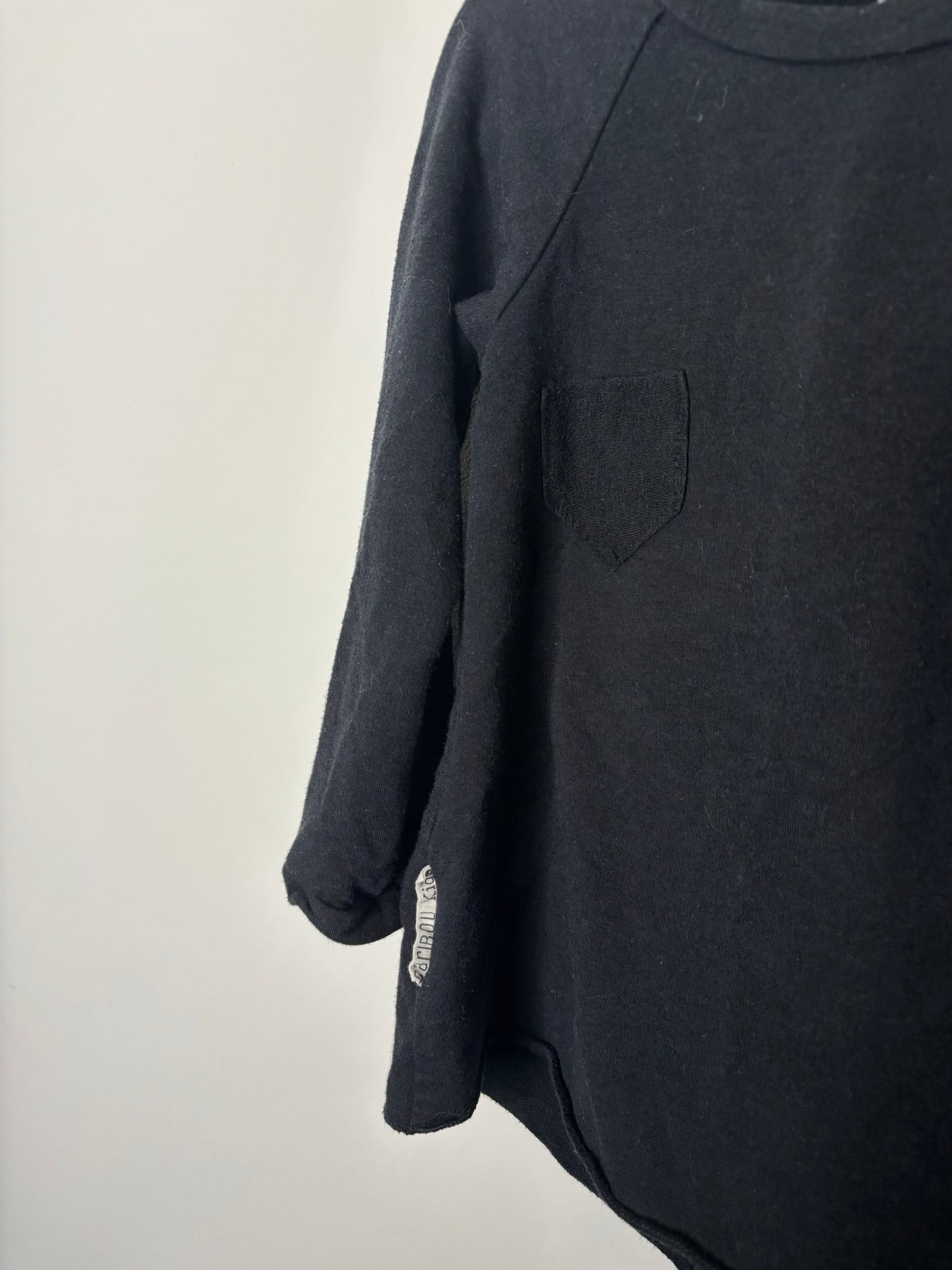 Black long sleeve tunic with a simple design, hanging on a grey hanger against a white brick wall.