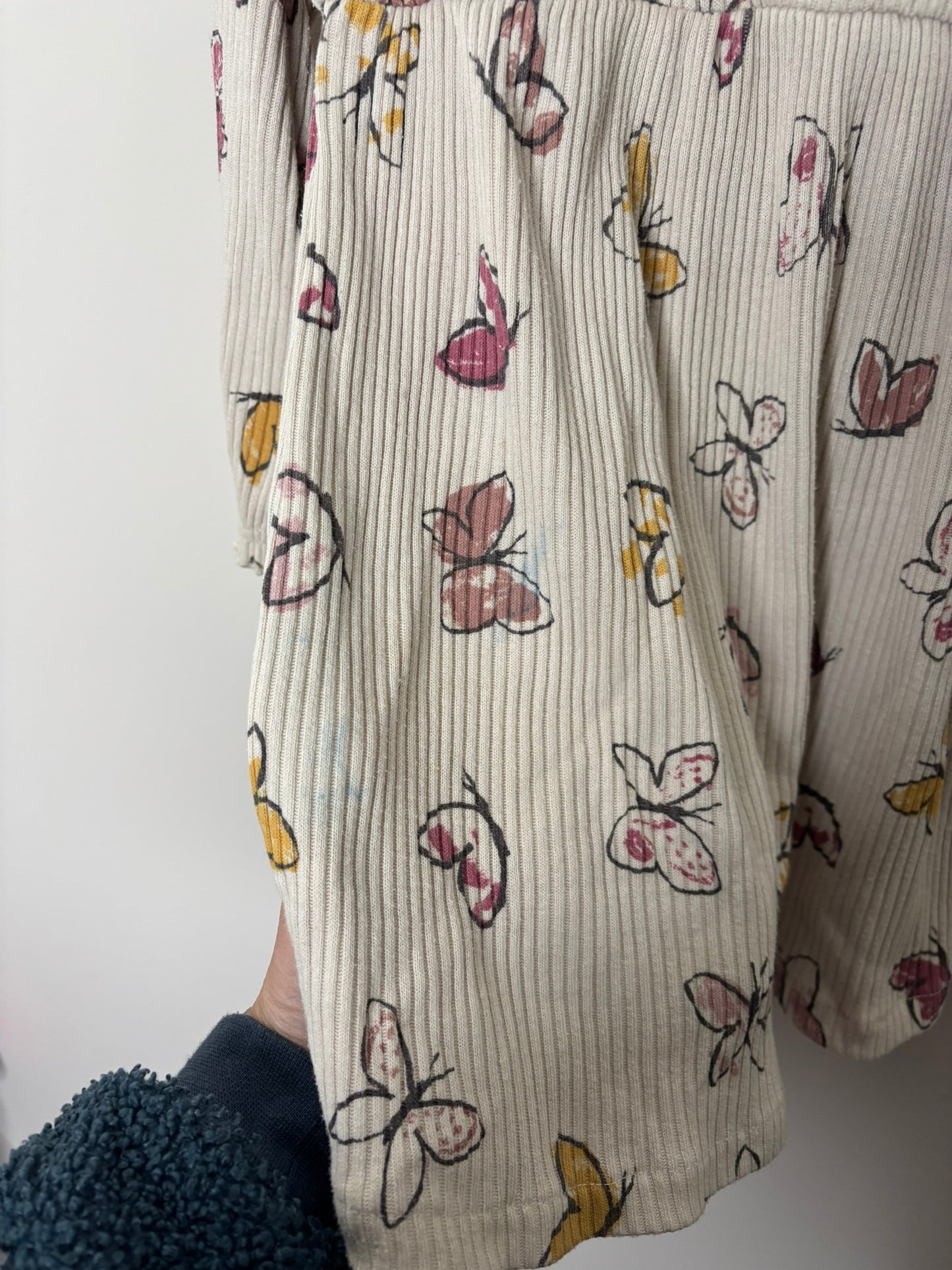 Cream long sleeve dress with a multicolored butterfly print, featuring buttons and a gathered waist, hanging against a white brick wall.