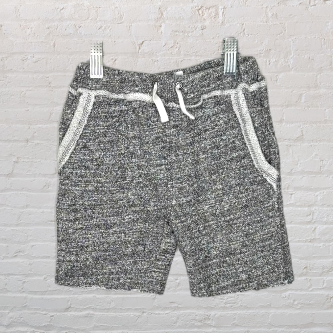 Grey knit shorts with white drawstring and pocket detailing, hung against a white brick background.