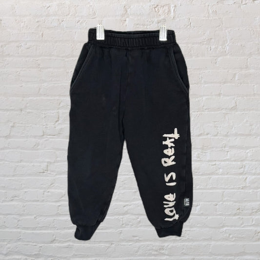 Black sweatpants with 'Love Is Real' text on one leg, displayed on hangers against a brick wall.