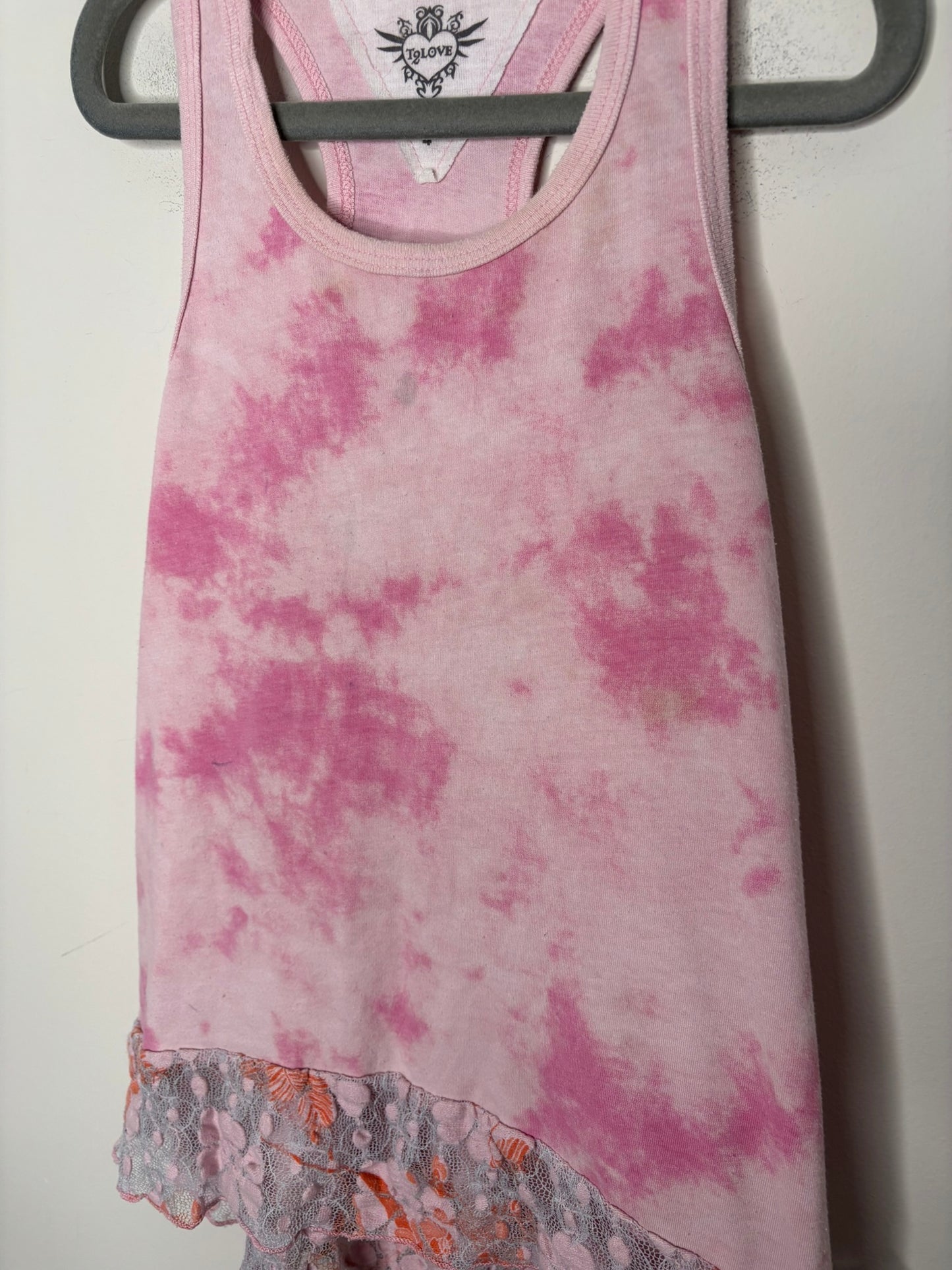 Pink tie-dye tank top with lace hem detail, hanging on a black hanger against a white brick wall.