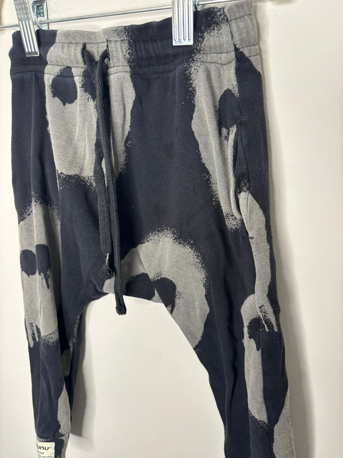 Black and gray drop crotch pants with skull print pattern and drawstring waist by NUNUNU, hanging on a white brick wall.