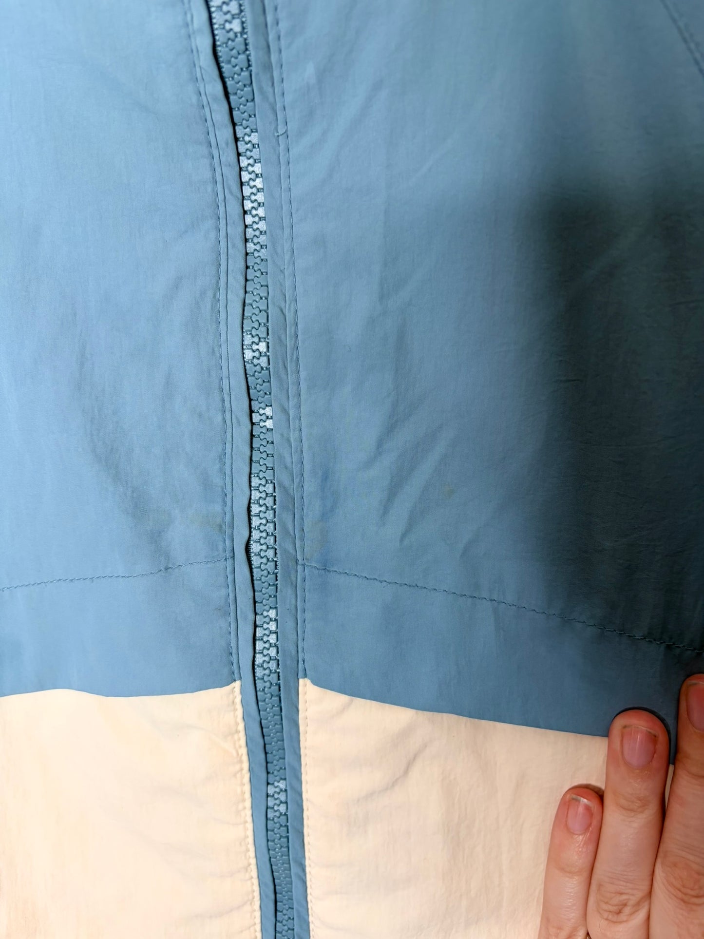 Blue and white hooded windbreaker jacket with front zipper and pockets, displayed on a hanger against a white brick wall.