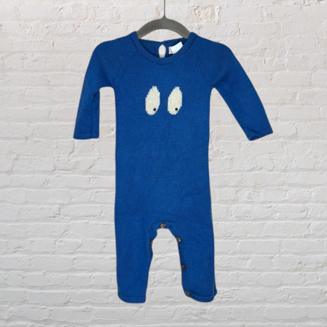 Blue knit onesie featuring a playful eye design on the chest, hanging on a dark hanger against a white brick wall.