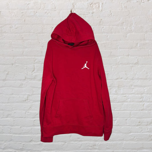 Red hoodie with a white Jumpman logo on the chest, featuring a front pocket, hanging against a brick wall.