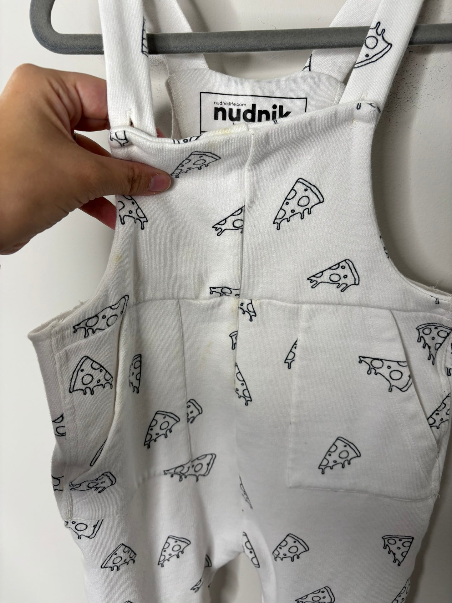 White overalls with black pizza print design, hanging on a dark hanger against a white brick wall.