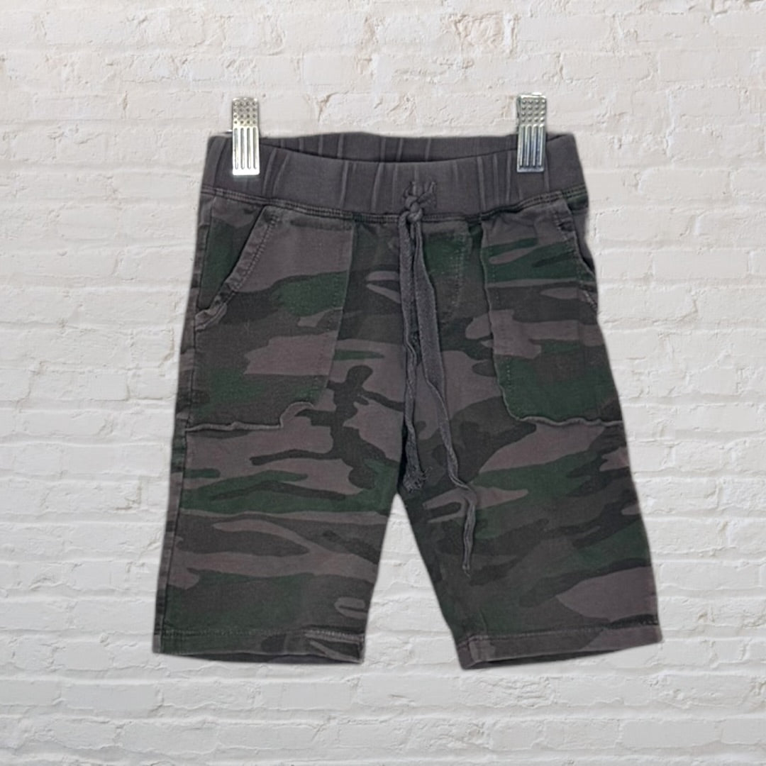 Camouflage print shorts with a drawstring waistband, displayed on a hanger against a white brick wall.