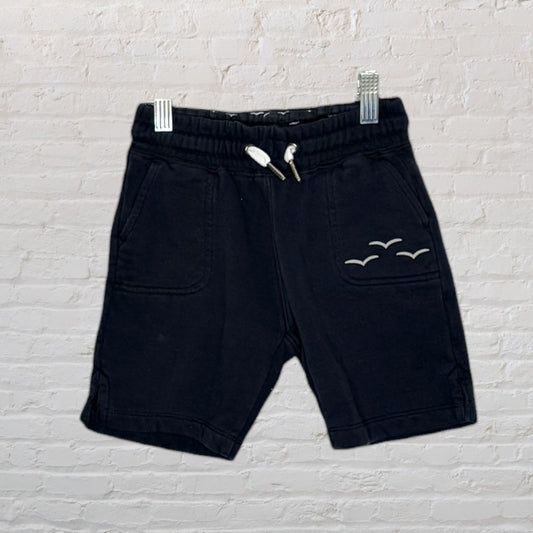 Black drawstring shorts with pocket and seagull embroidery, hanging on clips against a brick wall.