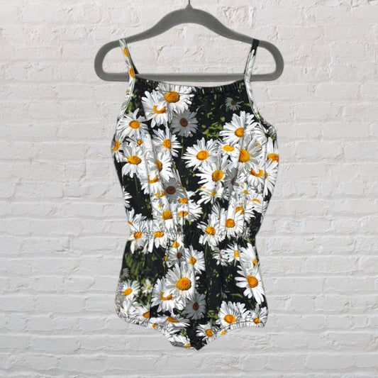 Women's black romper with white daisy floral print, featuring thin straps and elastic waist, hanging on a hanger against a white brick wall.