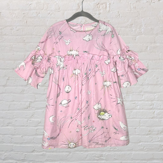 Pink dress with celestial illustrations and ruffled sleeves hanging against a white brick background.