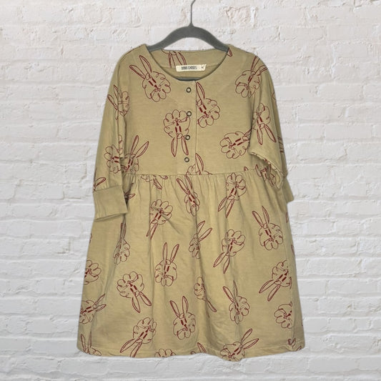 Beige long sleeve dress with red bunny print pattern by Bobo Choses, displayed on a hanger against a white brick wall.
