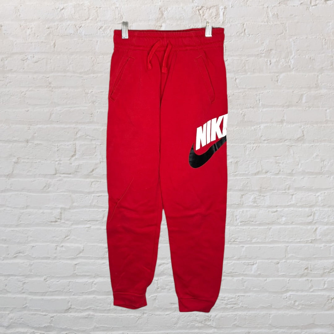 Red Nike sweatpants with a large white and black logo on the leg, displayed against a white brick wall.