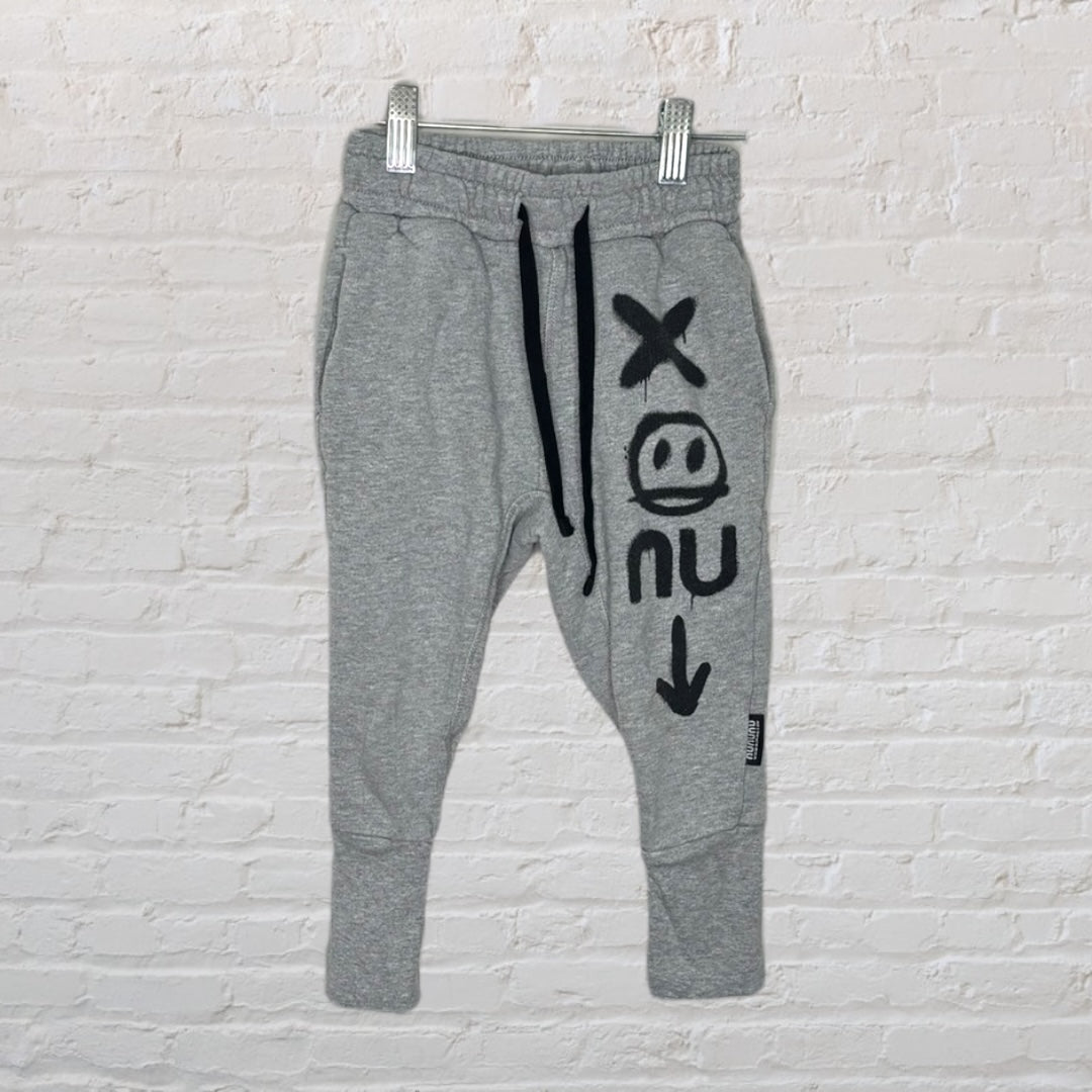 Grey NUNUNU sweatpants with black graphic designs, featuring an adjustable drawstring waist, hanging on a clip hanger.