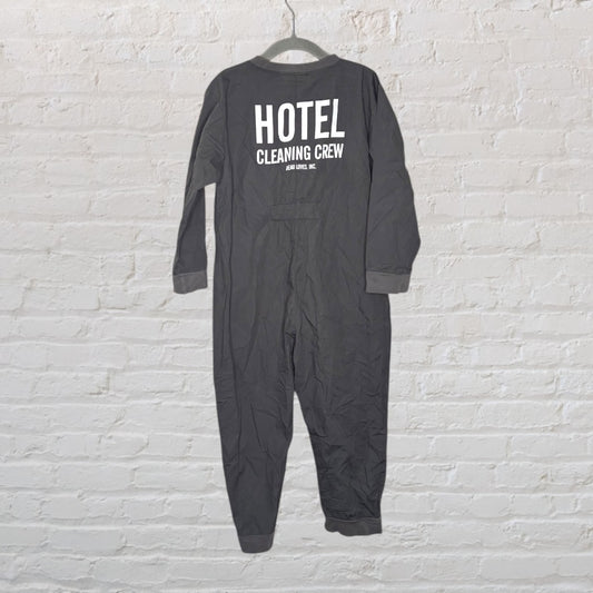 Dark grey coveralls with 'Hotel Cleaning Crew' printed on the back, displayed on a hanger against a white brick wall.