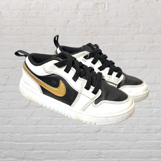 Black and white youth sneakers with a gold swoosh, featuring lace-up fronts, displayed against a white brick background.