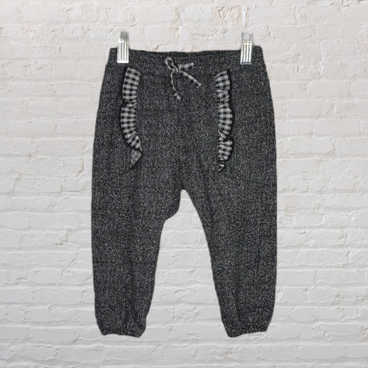 Charcoal knit pants with black and white gingham ruffle details, hanging against a white brick wall.