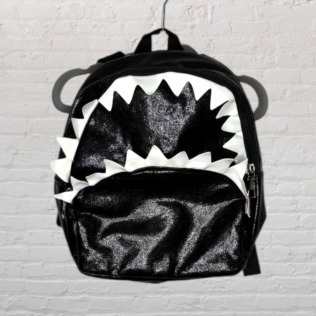 Black glittery shark-themed backpack with white jagged teeth design, hanging against a white brick wall.