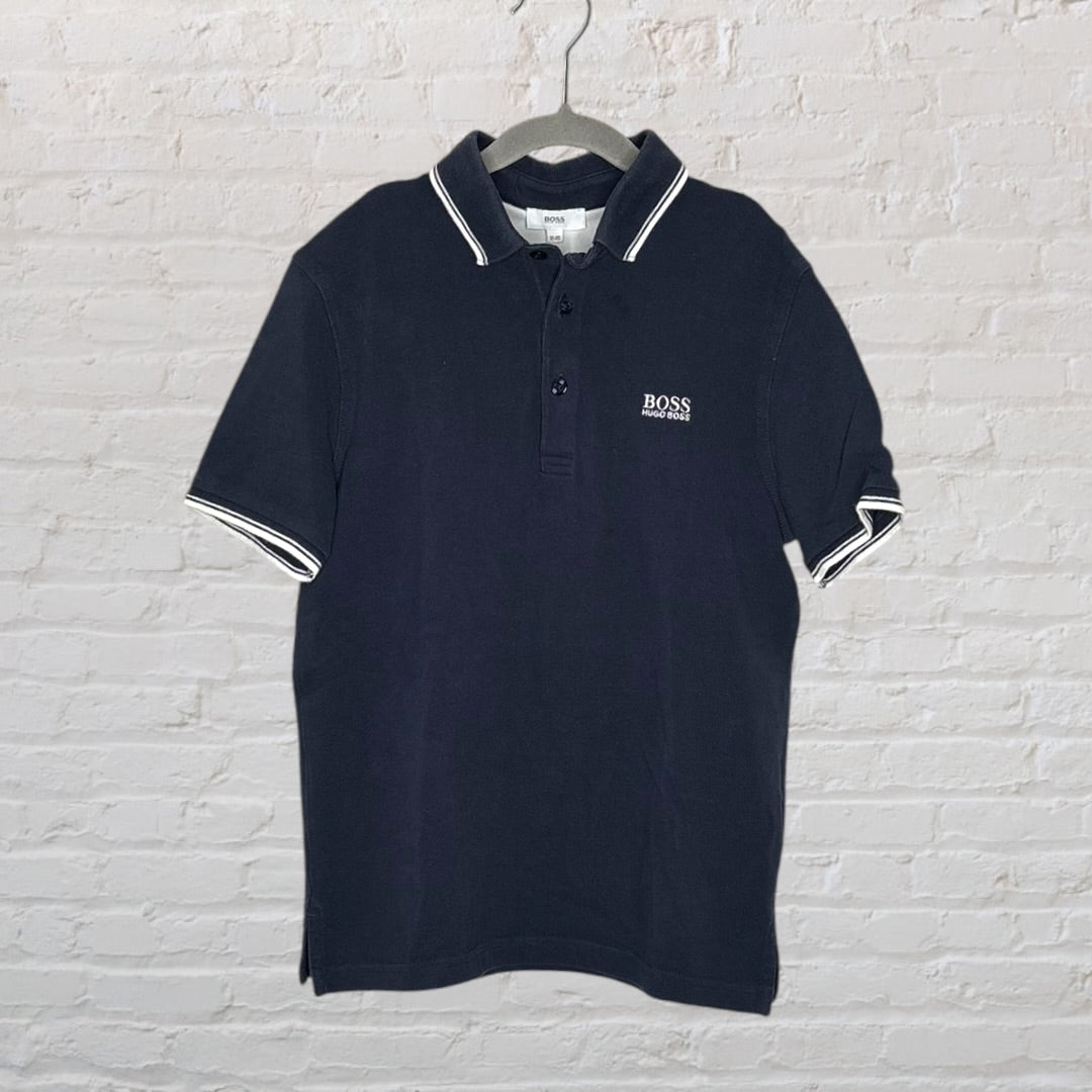 Navy blue polo shirt by BOSS Hugo Boss with white trim on collar and sleeves, hanging on a hanger.
