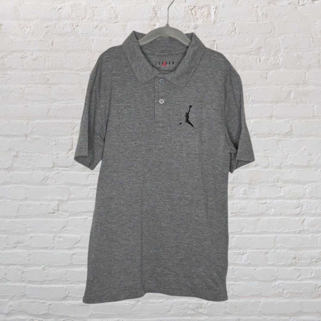 Grey Jordan polo shirt with embroidered Jumpman logo, hanging on a hanger against a white brick background.