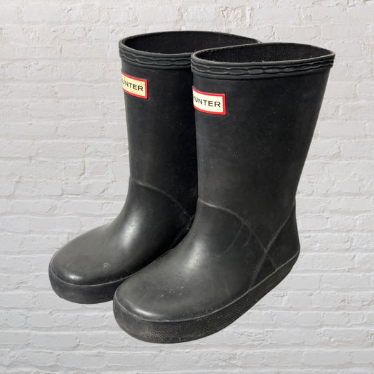 Pair of black Hunter rubber rain boots with red and white logo, set against a brick wall background.