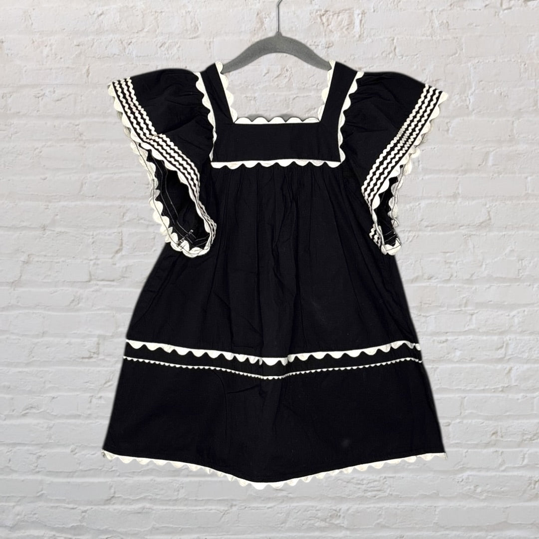 Black dress with white scalloped trims and flutter sleeves, hung on a hanger against a white brick wall.