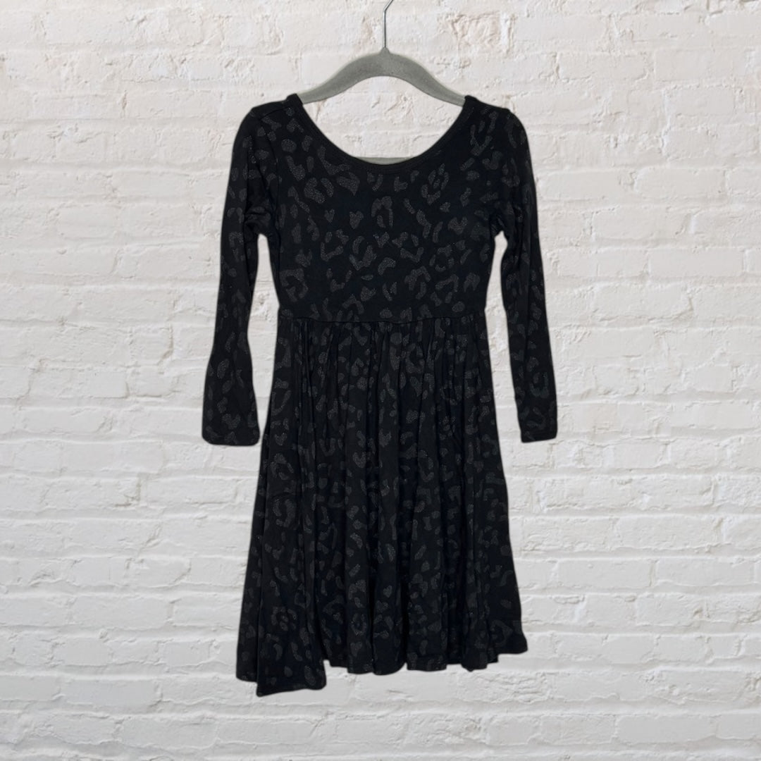 Black long-sleeve dress with subtle leopard print pattern, hanging on a hanger against a white brick wall.