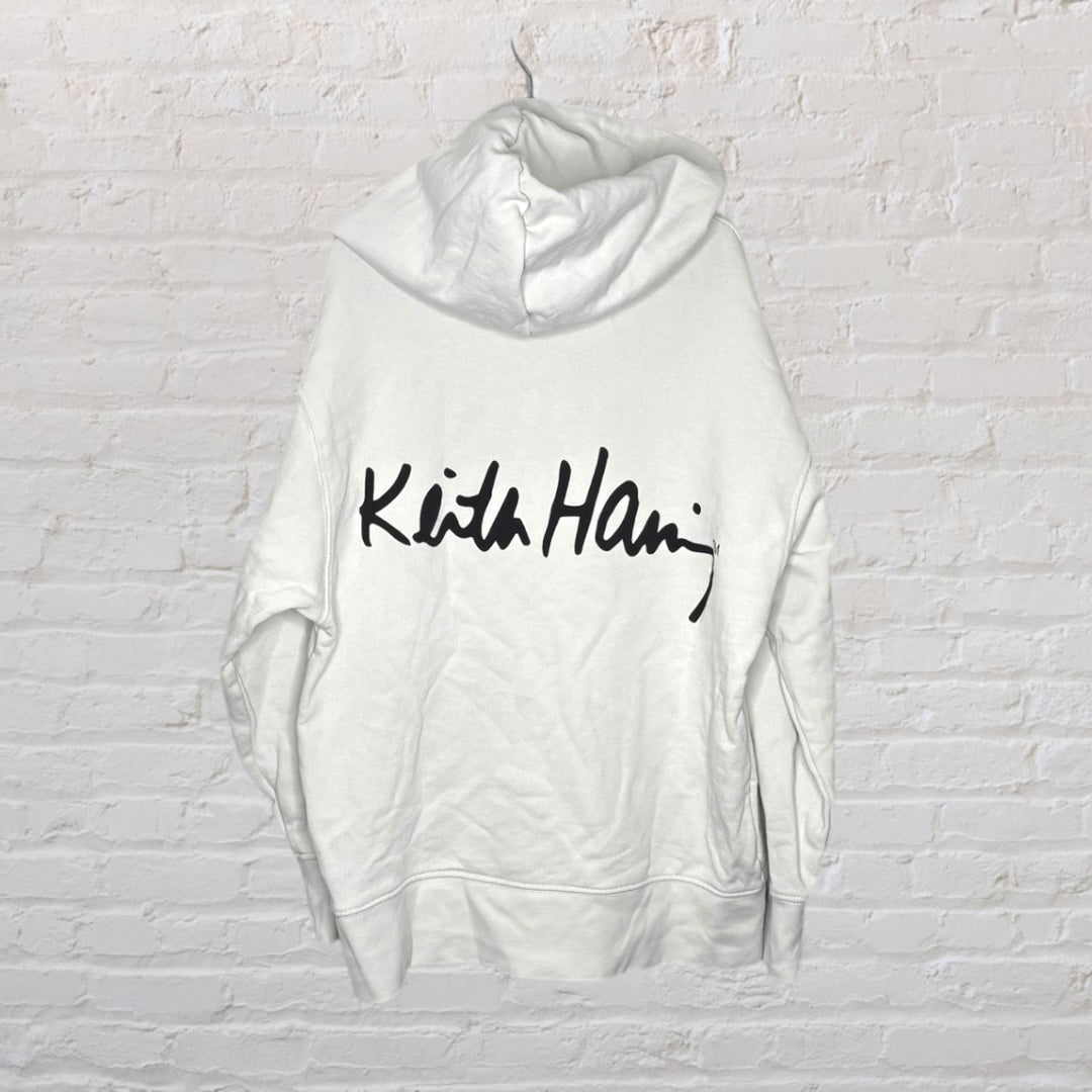 White hoodie with a Keith Haring style red figure on a skateboard graphic, hanging on a brick wall background.