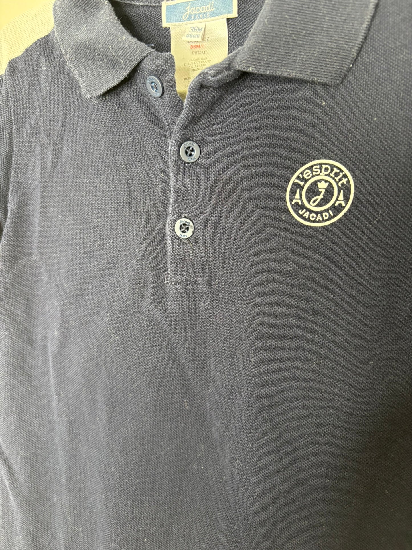 Black polo-style romper with white sleeves and a small embroidered logo, hanging on a hanger against a brick wall.