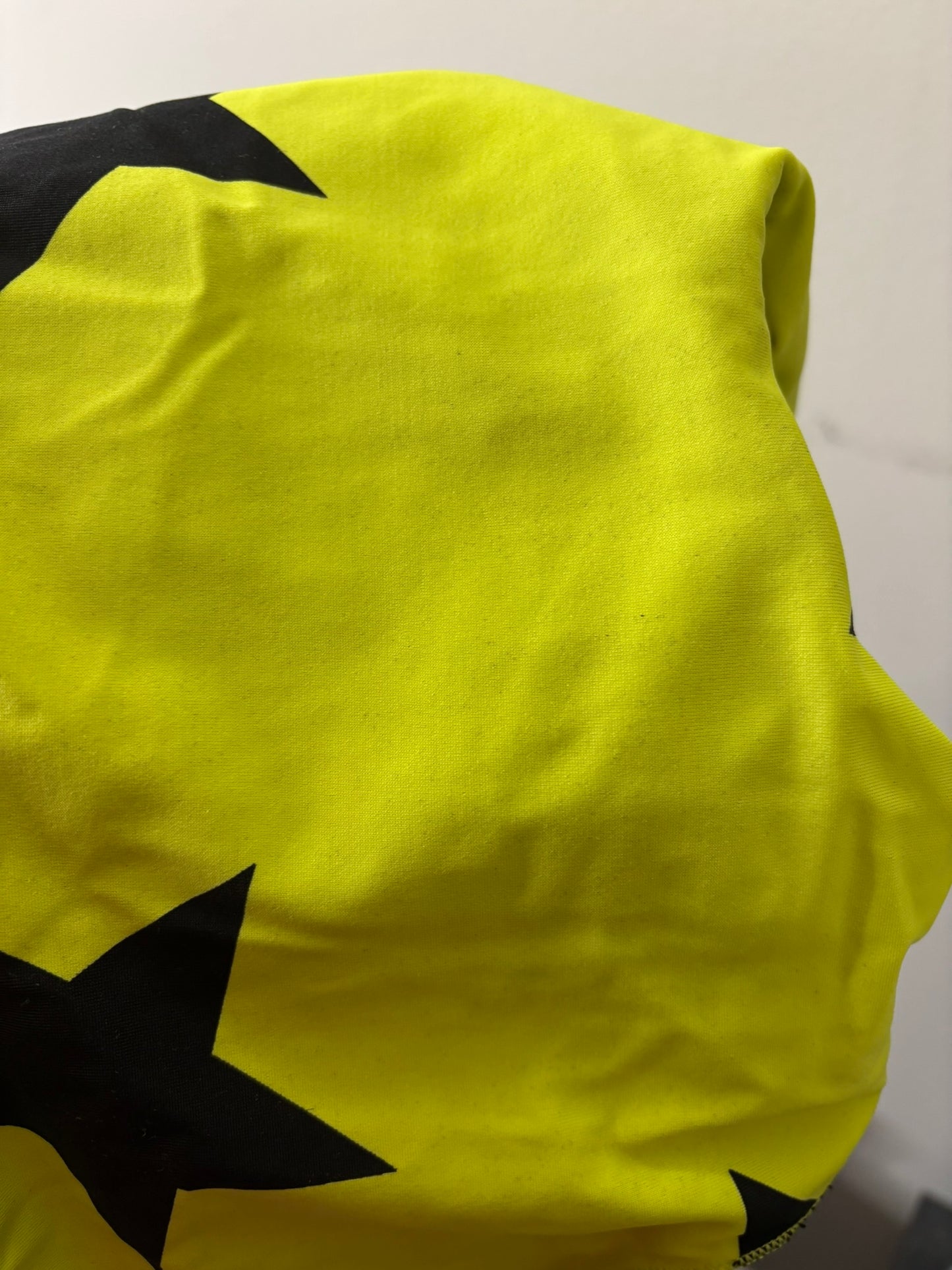 Neon yellow one-piece swimsuit with black star prints, hanging on a hanger against a white brick wall.