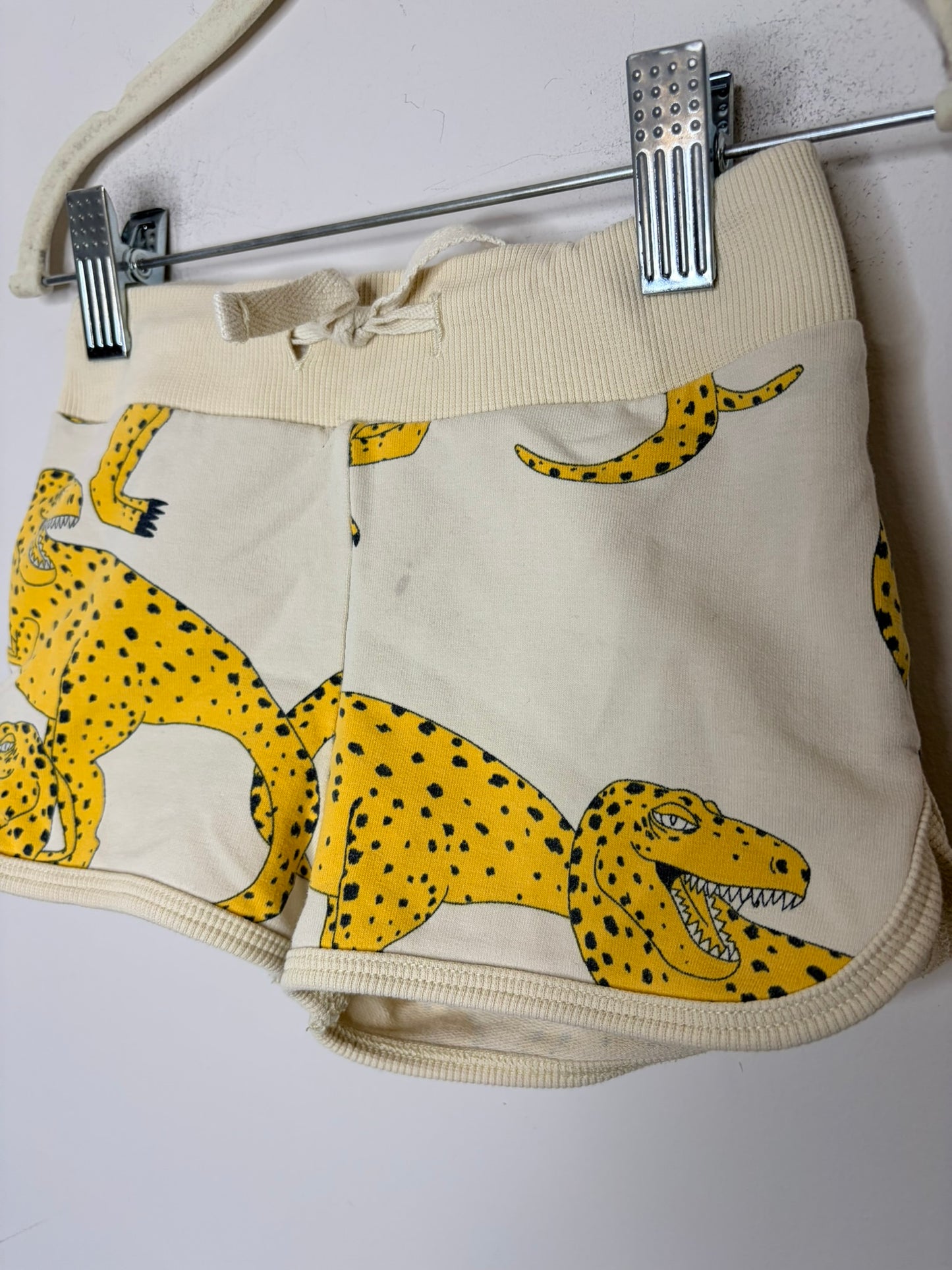 Beige shorts with yellow dinosaur print, displayed on a white brick background with clips.