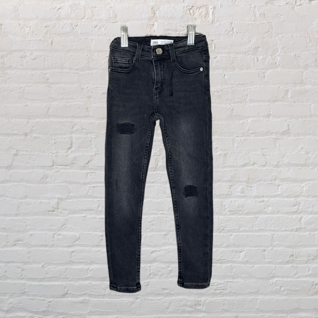 Black distressed skinny jeans for kids by Zara, displayed on hangers against a white brick wall.
