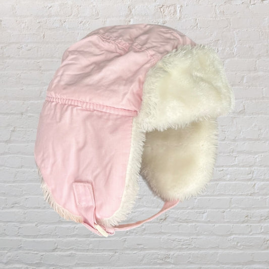 Pink trapper hat with white faux fur lining and ear flaps against a white brick background.