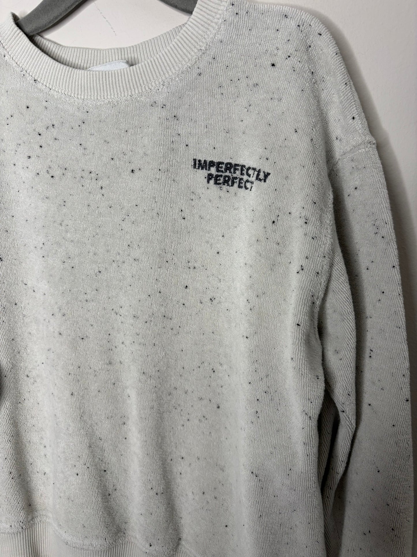 Light grey speckled sweatshirt with 'Imperfectly Perfect' text on the chest, displayed on a hanger against a brick wall.