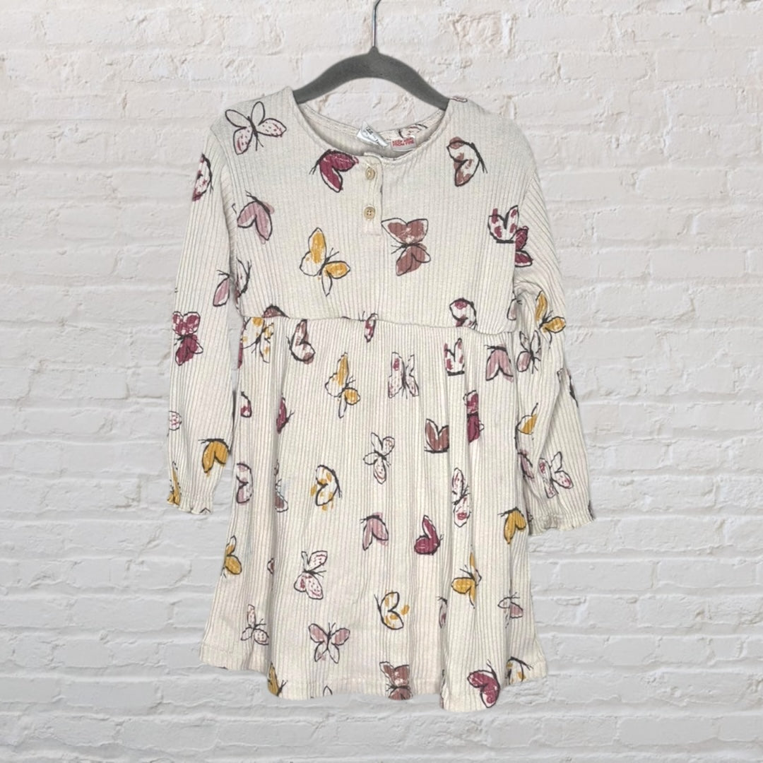 Cream long sleeve dress with a multicolored butterfly print, featuring buttons and a gathered waist, hanging against a white brick wall.