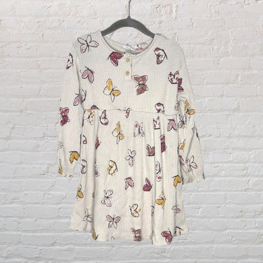 Cream long sleeve dress with a multicolored butterfly print, featuring buttons and a gathered waist, hanging against a white brick wall.