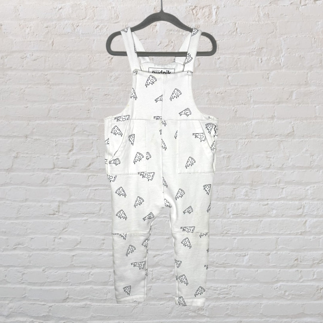 White overalls with black pizza print design, hanging on a dark hanger against a white brick wall.